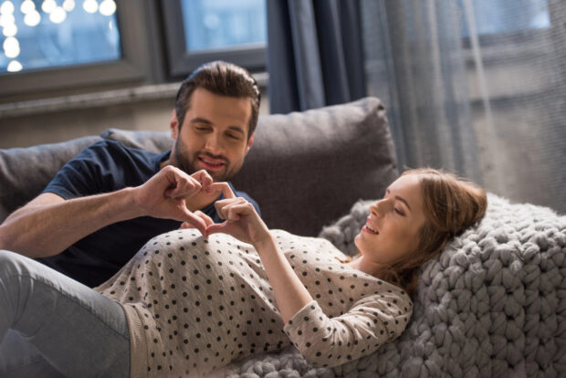 Schwangere Frau und Mann liegen auf einem Sofa und formen mit ihren Händen ein Herz.