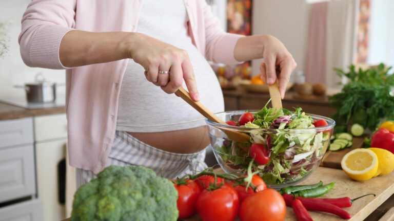 Schwangere Frau mischt frischen Salat in einer Glasschüssel mit Holzlöffeln in einer Küche.